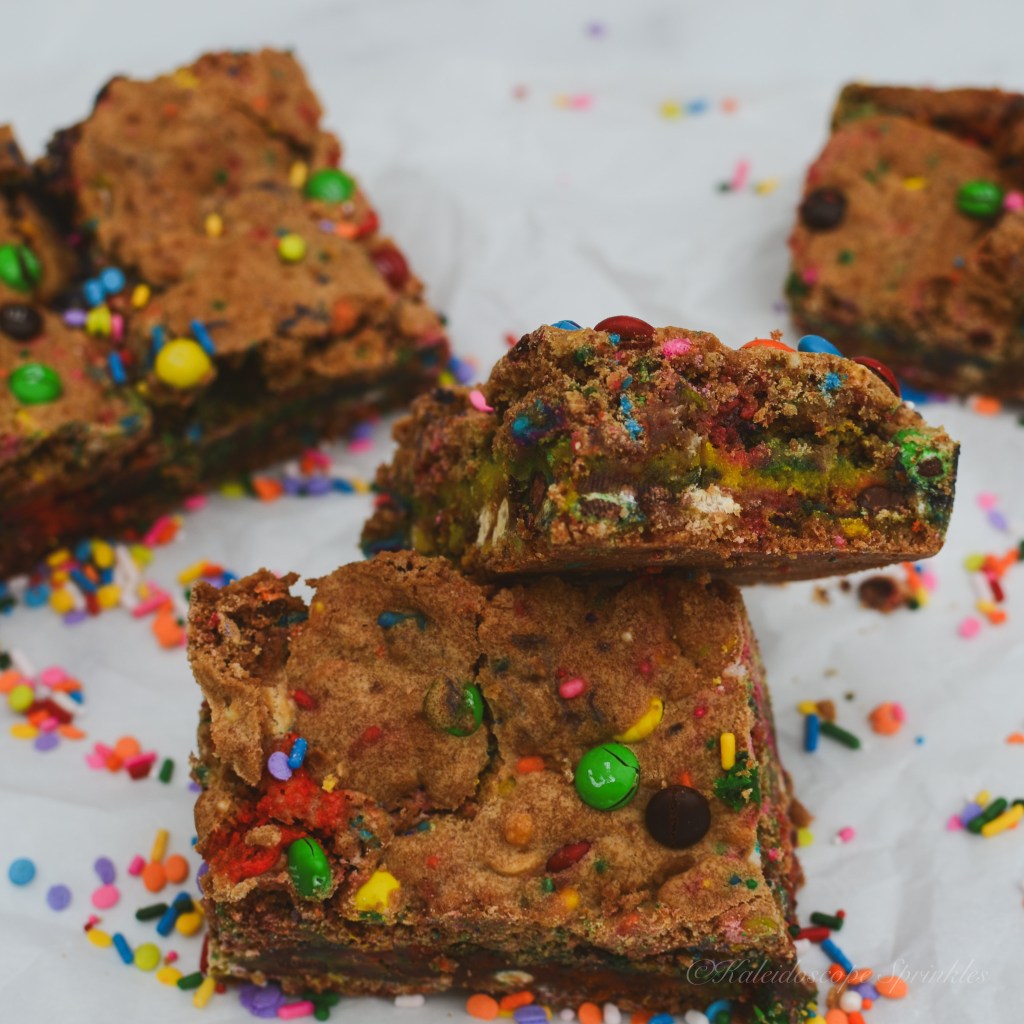 A colorful stack of Rainbow Blondies topped with vibrant sprinkles and surrounded by more scattered sprinkles, showcasing their chewy texture and festive appearance.