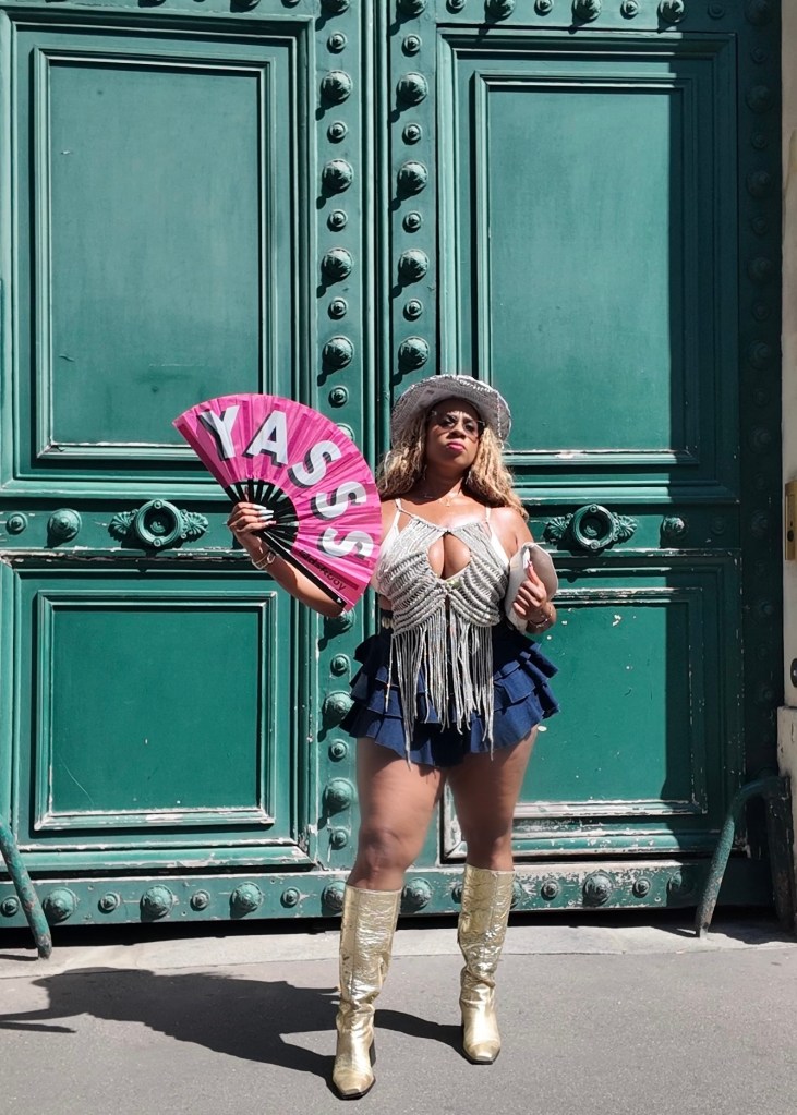 A person standing in front of a green door, wearing a sparkly silver top, a blue ruffled skirt, and metallic gold knee-high boots, holding a large pink fan that says 'YASS!'