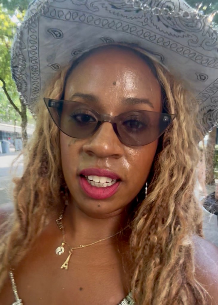 A close-up portrait of a person wearing a stylish cowboy hat with a bandana print and sunglasses, featuring wavy hair and pink lipstick.