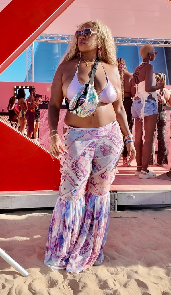 A person wearing custom-made Ribbon Corset Pants with a pastel international post card print, standing on a sandy beach at a festival.