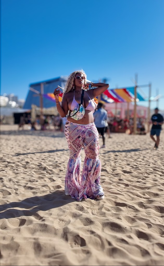 A person wearing a pink and purple bell-bottom pants with an international post card print, standing on the beach with a festive background and clear blue sky.