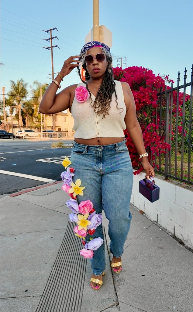 A person wearing a cropped white sweater and denim jeans decorated with faux flowers walking along a city street, showcasing a vibrant floral fashion statement.